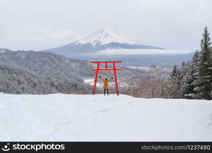 Asian woman tourist travel at Red Japanese Torii pole, Fuji mountain and snow in Kawaguchiko, Japan. Forest trees nature landscape background in winter season.