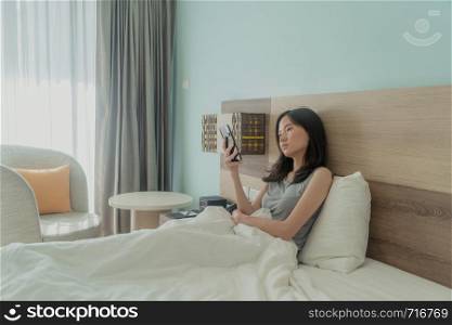 Asian woman using a mobile phone, thinking about problems and suffering from depression on bed in a modern bedroom with white blanket.