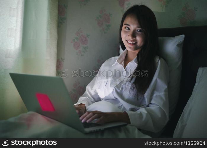 Asian woman using a notebook to work on night at the bed.She smiles and enjoys working at home.