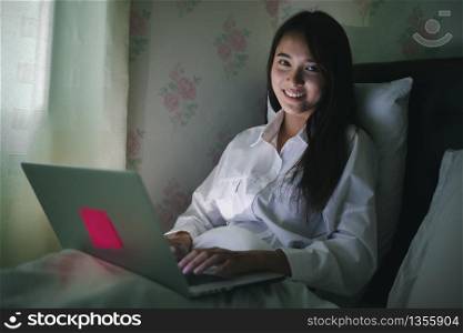 Asian woman using a notebook to work on night at the bed.She smiles and enjoys working at home.