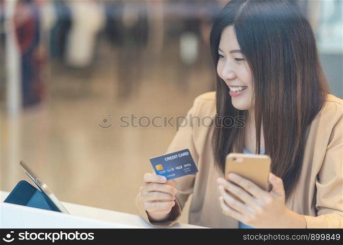 Asian woman using credit card with mobile phone for online shopping in department store over the clothes shop store background, technology money wallet and online payment concept, credit card mockup