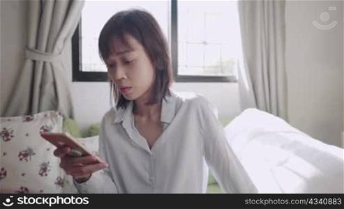 Asian woman using smartphone sitting down on couch at home living room, on bright sunny day, sending text message chatting, online communications technology, single female using dating application