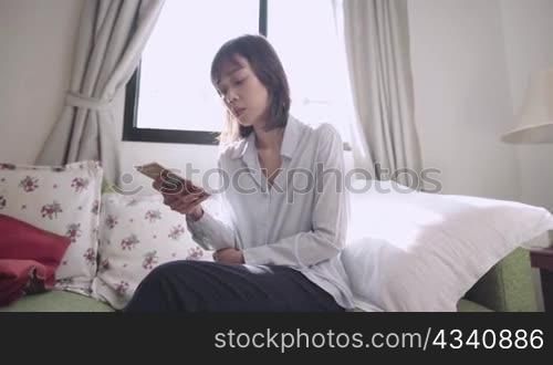 Asian woman using smartphone sitting down on couch at home living room, online dating application, sending email messaging online communications, house wife comfortable simple living, shopping online