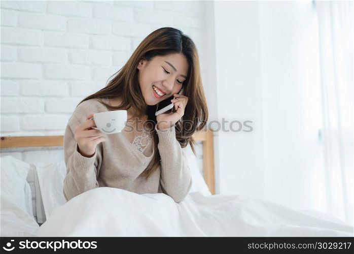 Asian woman using the smartphone on her bed while holding cup of. Asian woman using the smartphone on her bed while holding cup of coffee in the morning. Beautiful asian woman enjoying warm coffee and talking on telephone in her bedroom. lifestyle asia woman concept
