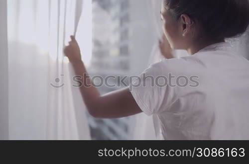 Asian woman wake up and open room curtain in the morning, view from high rise condo, with city street highway view on the outside, feeling fresh after good quality sleep wellness, view from behind