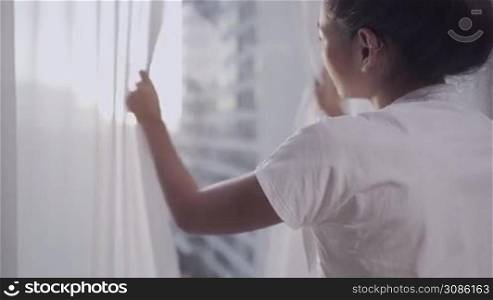 Asian woman wake up and open room curtain in the morning, view from high rise condo, with city street highway view on the outside, feeling fresh after good quality sleep wellness, view from behind