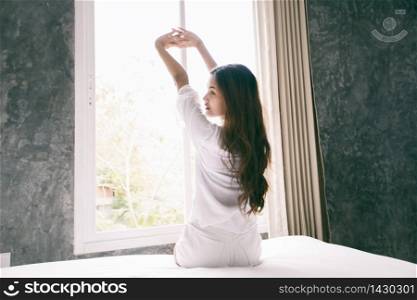 Asian woman waking up in her bed fully rested and open the curtains in the morning to get fresh air.