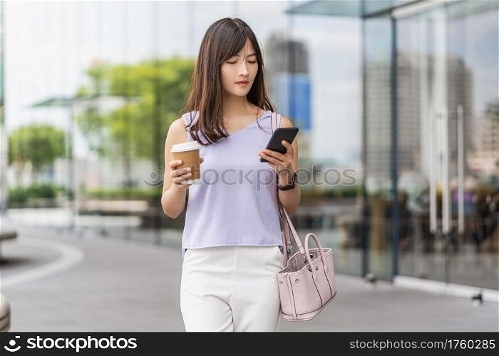 Asian woman walking in the city and using the mobile phone and holding a paper cup of coffee. department store shopping center, lifestyle and leisure, online shopping and cashless payment concept