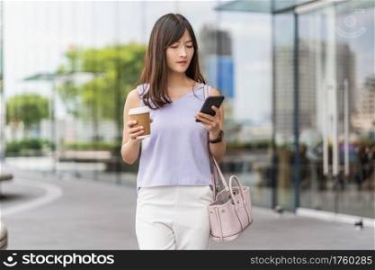Asian woman walking in the city and using the mobile phone and holding a paper cup of coffee. department store shopping center, lifestyle and leisure, online shopping and cashless payment concept