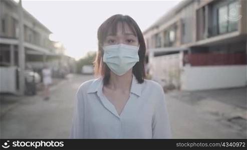 Asian woman wear face mask walking in front of house neighborhood street, Corona virus covid19 awareness caution, standing outdoor sunset social distancing concept, showing confidence by thumbs up