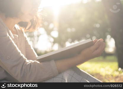 Asian woman wearing glasses is sitting reading a book on the grass while the sun sets in the park