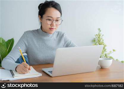 Asian woman wearing glasses is using laptop at home office.Portrait young cute student working on smart technology gadget