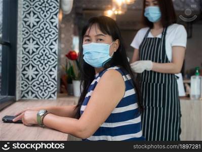 Asian woman wearing masks and sitting in a coffee shop and waitress in the mask are serving food. Concept of new lifestyle and disease prevention.