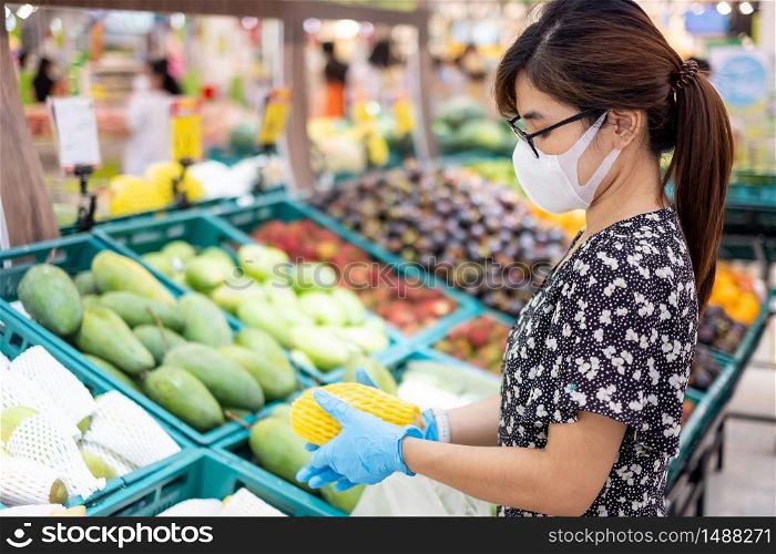 Asian woman wearing nitrile gloves and holding ripe mango fruit in supermarket or grocery, protect coronavirus inflection. Hygiene, new normal and life after covid-19 pandemic