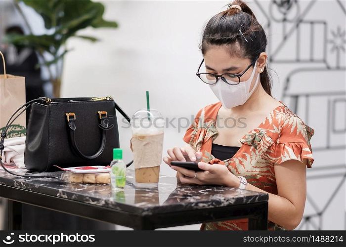 Asian woman wearing protective face mask and using smartphone during drinking coffee in restaurant, protect coronavirus inflection. social distancing, new normal and life after covid-19 pandemic