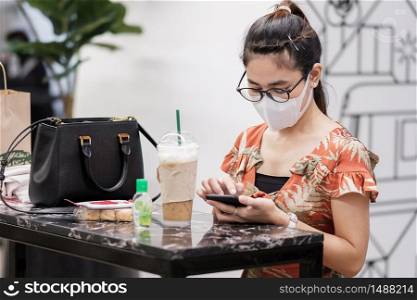 Asian woman wearing protective face mask and using smartphone during drinking coffee in restaurant, protect coronavirus inflection. social distancing, new normal and life after covid-19 pandemic