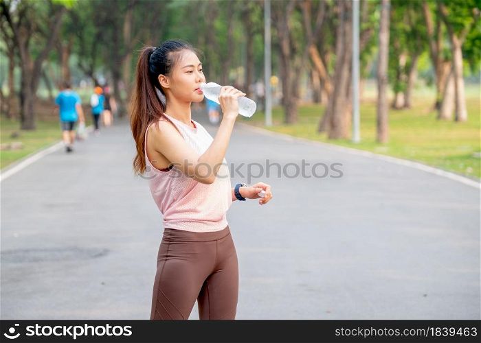 Asian woman with pink sportswear stand on road in park and drink water from bottle during exercise in the morning.