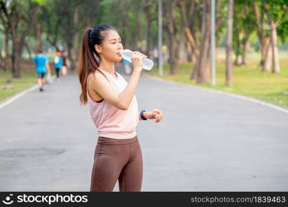 Asian woman with pink sportswear stand on road in park and drink water from bottle during exercise in the morning.