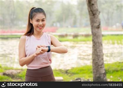 Asian woman with smiling touch her watch and look at camera with lake and flower field as background during her exercise in the morning.