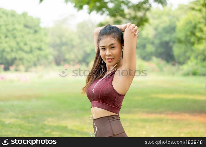Asian woman with sportswear do arm stretching and also look at camera after exercise in park or garden with morning light.