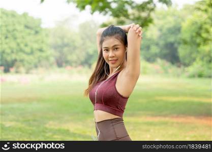 Asian woman with sportswear do arm stretching and also look at camera after exercise in park or garden with morning light.