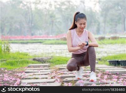 Asian woman with sportwear for jogging sit and look at her watch during exercise in garden or park in the morning with soft light.