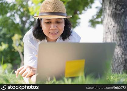 Asian woman work using a laptop while lying on green grass in a public park, lifestyle business concept