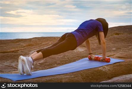 Asian woman workout in the morning at stone beach. Fit woman hand hold red dumbbell and push up on blue yoga mat for body strength. Weight training exercise for healthy lifestyle. Flat butt woman.