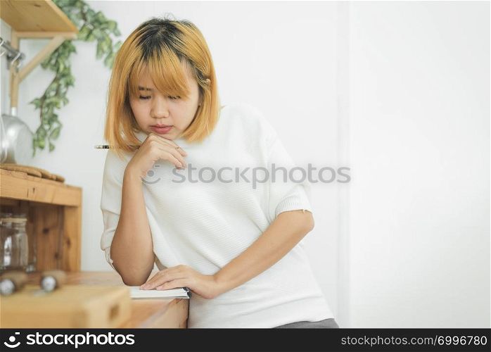Asian woman write shopping lists in notepad by pen on her kitchen counter at home and reading and tries to remember all ingredients to shopping in grocery. Lifestyle women relax at home concept.
