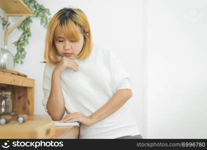 Asian woman write shopping lists in notepad by pen on her kitchen counter at home and reading and tries to remember all ingredients to shopping in grocery. Lifestyle women relax at home concept.