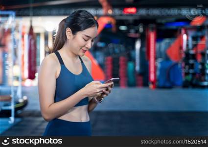 Asian Women are using mobile phones during exercise. She&rsquo;s standing, typing messages or chatting and smiling happily while staying during workouts with blur gym background and copy space