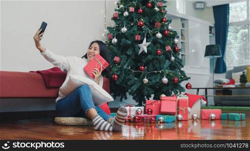 Asian women celebrate Christmas festival. Female teen relax happy holding Gift and using smartphone selfie with Christmas tree enjoy xmas winter holidays in living room at home.