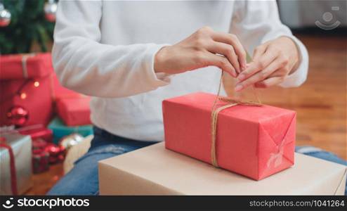 Asian women celebrate Christmas festival. Female teen wear sweater and Christmas hat relax happy wrapping gifts near Christmas tree enjoy xmas winter holidays together in living room at home.