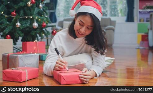 Asian women celebrate Christmas festival. Female teen wear sweater and santa hat relax happy write a wish on gift near Christmas tree enjoy xmas winter holidays together in living room at home.