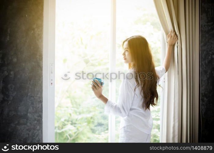 Asian women drinking coffee and wake up in her bed fully rested and open the curtains in the morning to get fresh air on sunshine