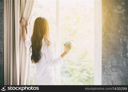 Asian women drinking coffee and wake up in her bed fully rested and open the curtains in the morning to get fresh air on sunshine