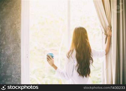 Asian women drinking coffee and waking up in her bed fully rested and open the curtains in the morning to get fresh air on sunshine