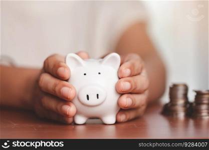 Asian women&rsquo;s hand holding a piggy bank with blurred coins and money on the wooden table for investment, business, finance and saving money concept.