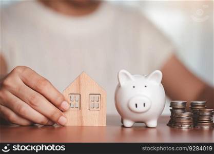 Asian women&rsquo;s hand holding a wooden house model with a saving piggy bank and a pile of coins and money on the wooden table for business, finance, saving money and property investment concept.