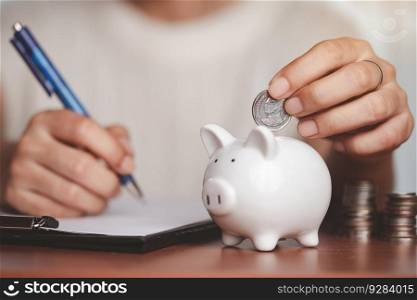 Asian women’s hand saving a coin into piggy bank, planning and writing information on paper with blurred coins and money on the wooden table for investment, business, finance and saving money concept.