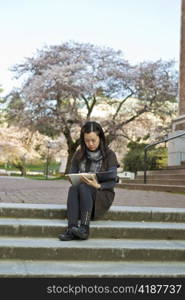 Asian women sitting down on steps while holding note book pads with cherry trees and blue sky in background