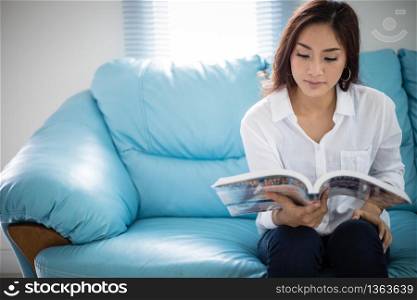 Asian women smiling and reading a book for relaxation on sofa at home