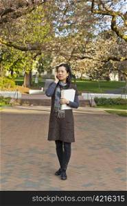 Asian women standing on sidewalk while holding note book pads with cherry trees and blue sky in background