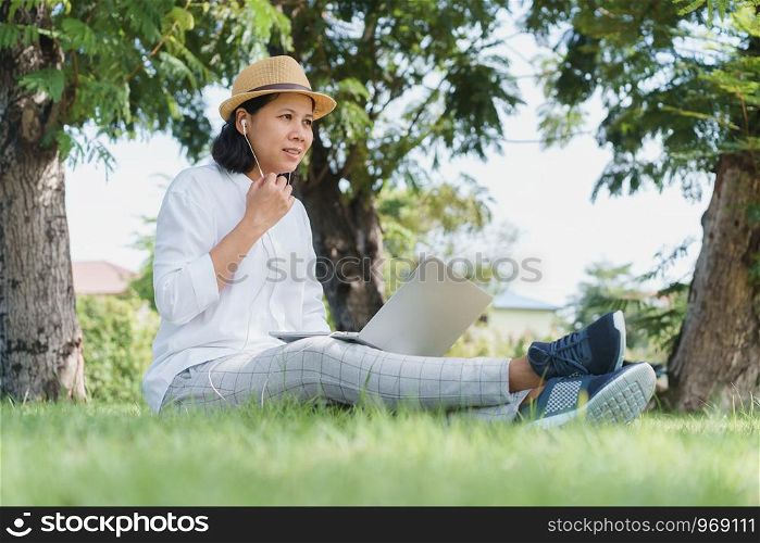 Asian women wear hat are using laptop with wired headphones while sitting on grass green In the public park