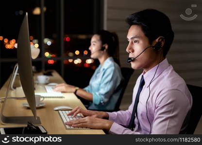 Asian young adult confidence operator man agent with headsets working in a call center at night Environment with his colleague team as customer service and technical support. Using as Late night Hard working 24 Hr. 7 Days concept.