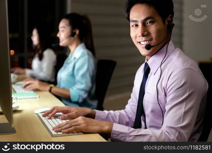 Asian young adult confidence operator man agent with headsets working in a call center at night Environment with his colleague team as customer service and technical support. Using as Late night Hard working 24 Hr. 7 Days concept.