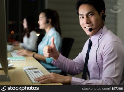 Asian young adult confidence operator man agent with headsets working in a call center at night Environment with his colleague team as customer service and technical support. Using as Late night Hard working 24 Hr. 7 Days concept.