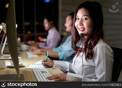 Asian young adult confidence operator woman agent with headsets working in a call center at night Environment with his colleague team as customer service and technical support. Using as Late night Hard working 24 Hr. 7 Days concept.