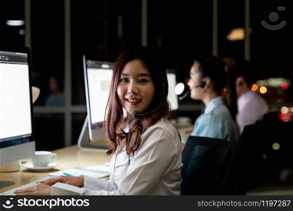Asian young adult confidence operator woman agent with headsets working in a call center at night Environment with his colleague team as customer service and technical support. Using as Late night Hard working 24 Hr. 7 Days concept.