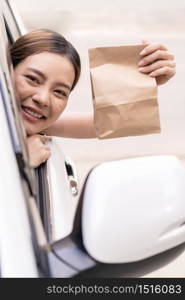 Asian Young adult in car holding disposable bag for take out food from drive thru service restaurant. Drive thru is new normal and popular service after coronavirus covid-19 pandemic.